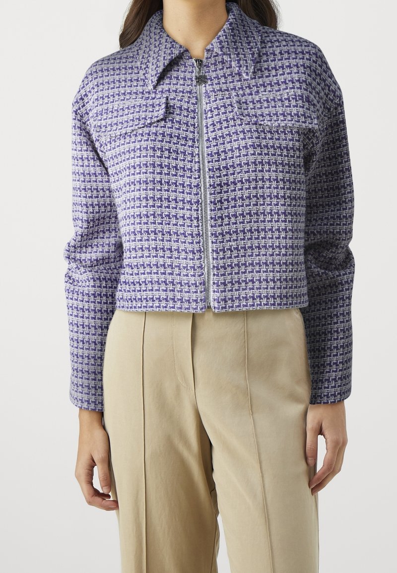 Femme portant une veste courte à carreaux violets et blancs zippée, avec deux poches poitrine, et un pantalon beige taille haute.