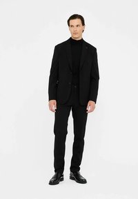 Sort jakkesæt med en tætsiddende blazer og bukser, kombineret med en sort rullekrave. Har et strømlinet design, glat tekstur og skinnende sorte sko.