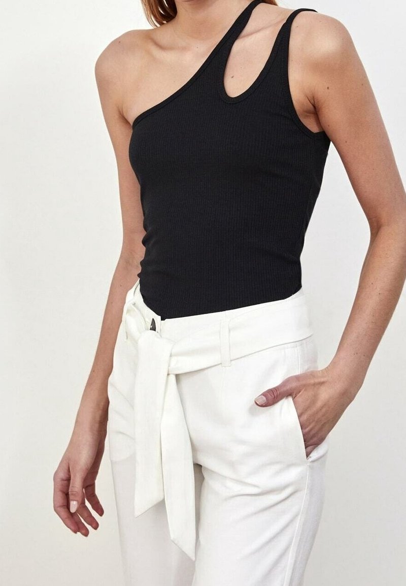 Femme portant un haut noir asymétrique et un pantalon blanc taille haute avec une ceinture nouée, debout avec une main dans la poche.