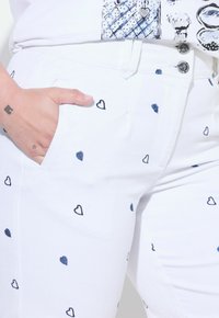 Pantalones blancos con pequeños estampados de corazones en azul marino, con bolsillos frontales y detalles de botones plateados. Tejido suave con una textura lisa.