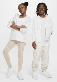 Dos niños llevan ropa deportiva: una chaqueta blanca con cremallera, Leggings beis claros y zapatillas blancas. El niño tiene el pelo largo y negro; la niña lleva un moño.