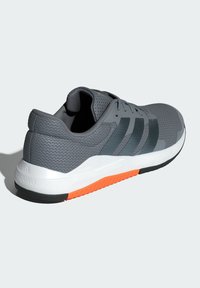 Zapatilla deportiva gris con parte superior de malla, superposiciones texturizadas y tres rayas oscuras. Media suela blanca con suela de tracción naranja.