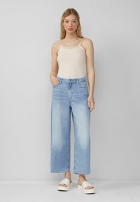 Helles beige Trägershirt mit Spitzenbesatz, kombiniert mit hochtaillierten, hellblauen Weitbeinjeans und weißen Plateausandalen. Einfaches, klares Design.