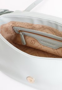 Vaaleansininen nahkalaukku, jossa on vetoketjullinen tasku ja monogrammilla varustettu vaaleanbeige vuori, jossa on erilaisia kuvioita. Hienovaraisia kultaisia varusteita.