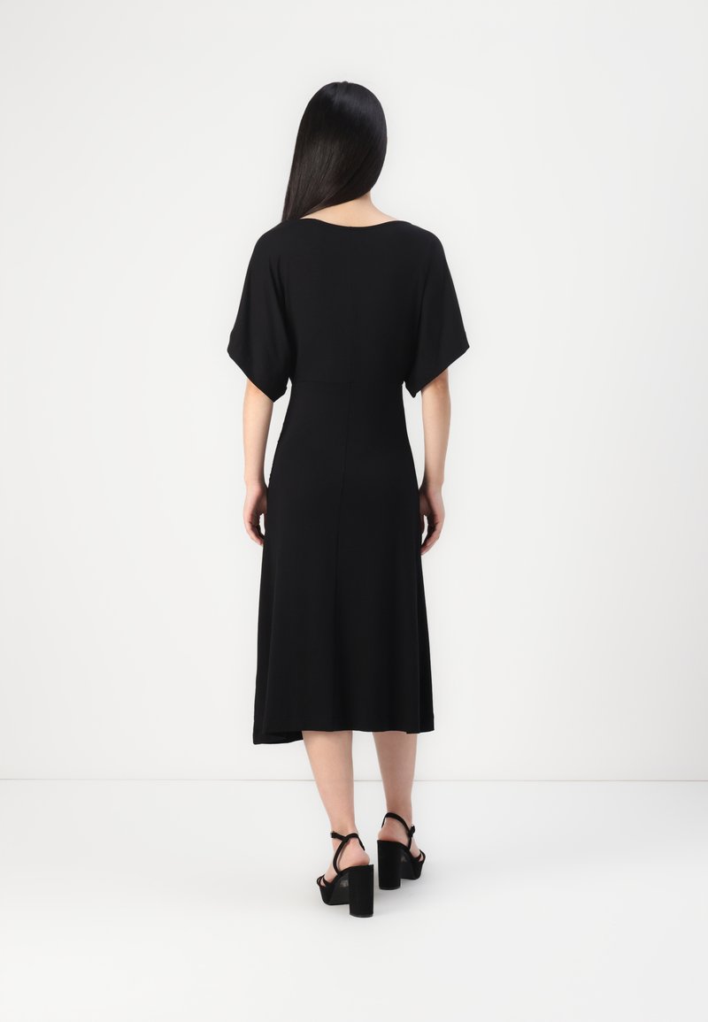 Zwarte, knielange jurk met een losse pasvorm, vleermuismouwen en een zachte textuur. Draag met zwarte hoge hakken. Achteraanzicht weergegeven.