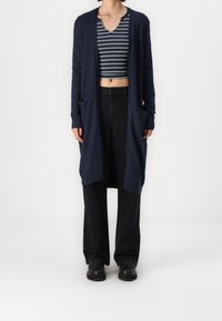 Cárdigan largo de color navy con una textura suave, de frente abierto y con bolsillos laterales, combinado con un top de rayas corto y pantalones negros de cintura alta.