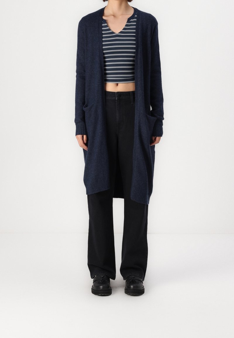 Cárdigan largo de color navy con una textura suave, de frente abierto y con bolsillos laterales, combinado con un top de rayas corto y pantalones negros de cintura alta.