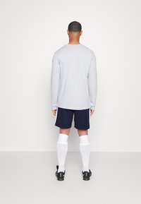 Camisa de manga larga gris, pantalones cortos deportivos azules marinos, calcetas blancas hasta la rodilla y zapatos negros, vistos desde la parte posterior sobre un fondo liso.