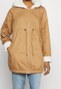 Osoba v krémovém zipovaném kabátu s bílým fleecem v kapuci a na manžetách, s rukama v kapsách, ve světle modrých džínách.