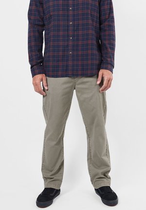 Homme portant une chemise à carreaux bleu foncé et rouge boutonnée, un pantalon marron clair et des baskets noires, debout devant un fond uni.