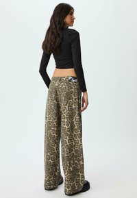 Donna con lunghi capelli scuri che indossa un crop top nero a maniche lunghe e pantaloni a gamba larga con stampa leopardata, in piedi di lato su uno sfondo neutro.