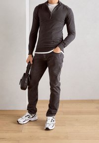 Maglione grigio scuro aderente con zip sopra una camicia bianca, abbinato a jeans scuri e sneakers bianche, mentre tiene una piccola borsa nera. Sfondo con pavimento in legno.