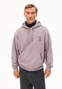 Uomo di mezza età con capelli corti e barba, che indossa una felpa lavanda sopra un dolcevita nero e jeans scuri, in piedi contro uno sfondo bianco.