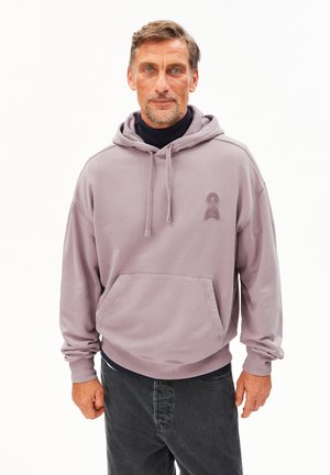 Middelbare leeftijd man met kort haar en een baard, gekleed in een lavendelkleurige hoodie over een zwarte turtleneck en donkere jeans, staande tegen een witte achtergrond.
