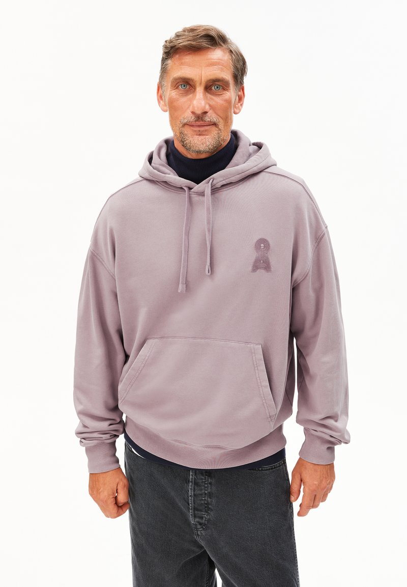 Middelbare leeftijd man met kort haar en een baard, gekleed in een lavendelkleurige hoodie over een zwarte turtleneck en donkere jeans, staande tegen een witte achtergrond.