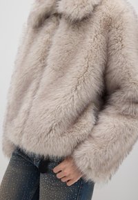 Korte nepbontjas in licht beige, met een kraag, brede mouwen en een zachte textuur. Draagt men over denim met glinsterende details.