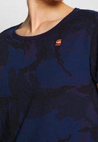 Laivansininen lyhythihainen t-paita, jossa on hillitty kamppailupainatus, pyöreä kaula-aukko ja pieni oranssi brodeerattu logo rinnassa.