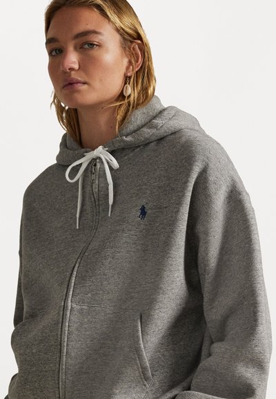 Šedá mikina na zips z mäkkého materiálu, ktorá obsahuje biele šnúrky, predné vrecko a malé modré logo na ľavej hrudi.