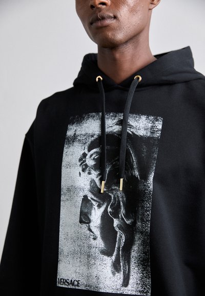Sudadera negra con un diseño gráfico que presenta una escultura clásica en monocromo. Los cordones negros con puntas doradas realzan el diseño.