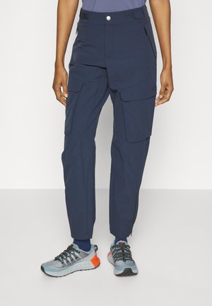 Pantalon cargo bleu marine avec fermeture éclair et poches à rabat porté avec des chaussures de course bleues et orange et des chaussettes bleues sur une personne debout.