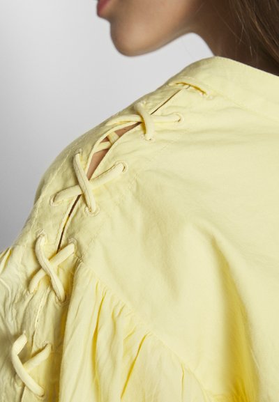 Chemise jaune clair avec un détail de laçage sur l'épaule, en coton doux, texture froncée et encolure large.