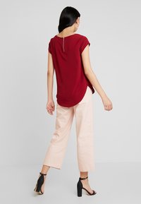 Bordowa bluzka z krótkim rękawem, okrągłym dekoltem i suwakiem z tyłu, zestawiona z bladoróżowymi szerokimi spodniami i czarnymi sandałami na obcasie.