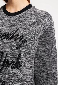 Grau gestrickter Pullover mit einer strukturierten Oberfläche, schwarzem Rundhalsausschnitt und gesticktem "Superdry New York"-Logo in schwarfem Faden.
