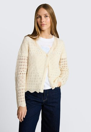 Young woman with long hair wearing a cream knit cardigan over a white t-shirt and dark blue jeans, with one hand in pocket.