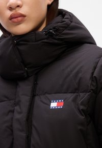 Casaco acolchoado preto com colarinho alto, fecho em zíper à frente e logótipo bordado "TOMMY JEANS" em vermelho, branco e azul no lado esquerdo do peito. Textura suave.