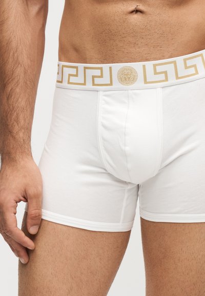 Torse masculin portant un boxer blanc avec une ceinture dorée à motif et logo Méduse, main reposant près de la cuisse, teint de peau brun moyen.