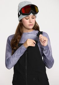 Salopette noire avec bretelles réglables, portée sur un t-shirt à manches longues violet clair. Bonnet en tricot gris et lunettes de ski colorées posées sur le front.