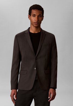 Joven con una blazer gris oscuro sobre una camisa negra, posando frente a un fondo liso gris claro.