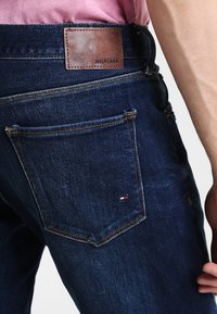 Jeans i mörk denim med läderetikett "HILFIGER", guldsömmar och en bakficka med en liten röd, vit och blå logotyp.