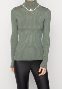 Long-sleeve, ribbed turtleneck sweater in muted green, featuring a fitted design. The model wears layered decorative necklaces.
