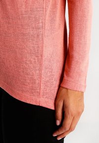 Langärmliger, korallenfarbener Strickpullover mit strukturiertem Stoff, seitlichen Schlitzen und leicht geschwungenem Saum. Nahaufnahme einer Hand, die den Stoff berührt.