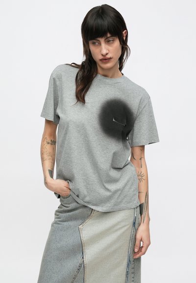 Tumehalli naine, kes kannab halli T-särki musta spritiga kaetud ringiga rinnatasku kohal ja kahetoonilist teksaseelikut.