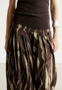 Vrouw in een donkerbruin mouwloos top en een lange, geplooide rok met gouden en bruine abstracte strepen, geaccessoireerd met een brede zilveren armband.