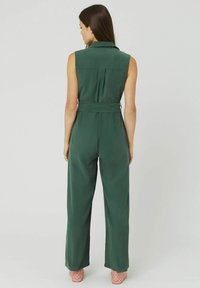 Combinaison sans manches verte avec une taille cintrée, des jambes larges et des poches arrière. Le tissu a une texture lisse et une couleur unie.