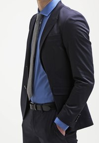 Tmavě modré sáčko ve vojenském stylu přes světle modrou košili, šedou kravatu a tmavé kalhoty. Má elegantní design, hladkou texturu a jemné detaily kapes.