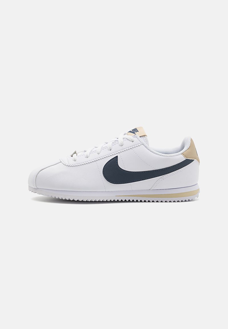 Hvite joggesko med marineblå swoosh, beige hælakkent, teksturert gummisåle og klassisk snøring, laget av glatt syntetisk materiale.