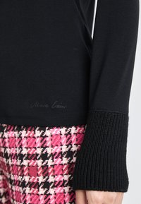Schwarzes Langarmoberteil mit gerippten Bündchen und einem dezenten Logo, kombiniert mit einem gemusterten Stoff in pink und schwarz im Hahnentrittmuster.