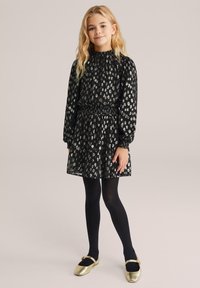 Crna haljina s nabranim dekolteom i dugim rukavima, sadrži srebrni uzorak leoparda. Suknja ima slojevite volančiće. Uparena je s crnim najlonkama i zlatnim cipelama.