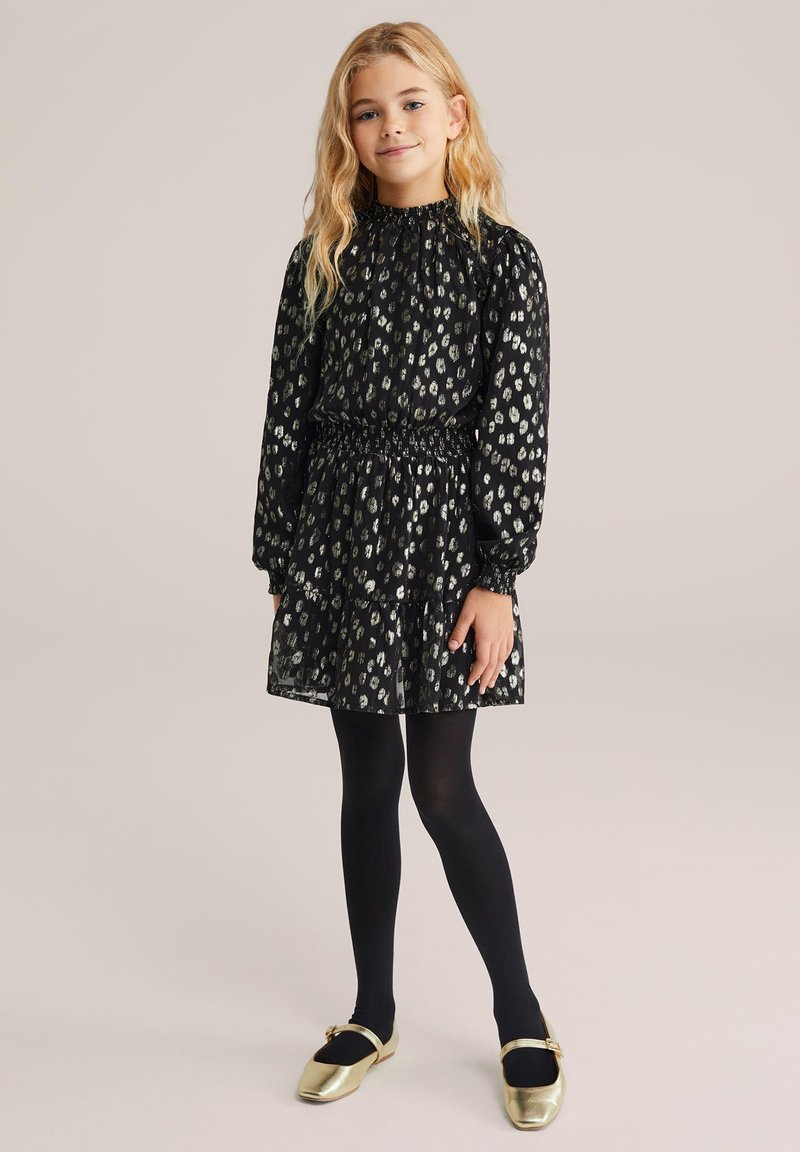 Crna haljina s nabranim dekolteom i dugim rukavima, sadrži srebrni uzorak leoparda. Suknja ima slojevite volančiće. Uparena je s crnim najlonkama i zlatnim cipelama.