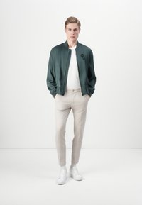 Veste bomber en tissu vert foncé avec un logo noir, associée à un pantalon beige clair et des baskets blanches. Design simple et ajusté avec une texture lisse.