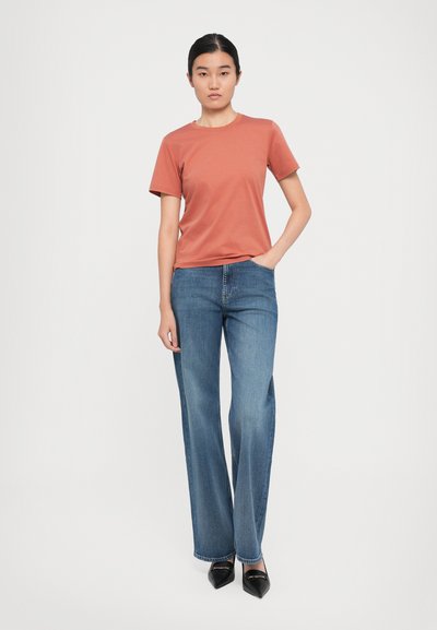 Ung kvinna står med ena handen i fickan, iförd en rostfärgad kortärmad skjorta, blå vida jeans och svarta spetsiga skor.