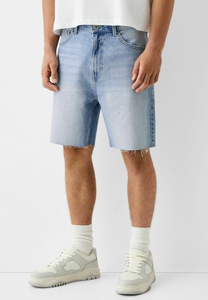 Shorts en jean bleu clair avec ourlet effiloché, style cinq poches et coupe décontractée. Associés à des baskets grises et des chaussettes blanches montantes.