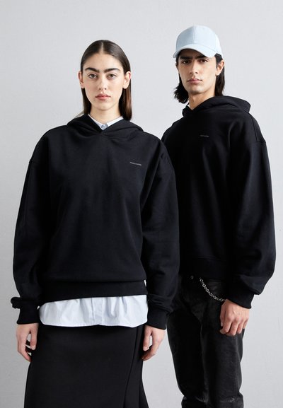 Un sweat à capuche noir avec une poche avant, une coupe oversize et des poignets côtelés. Porté par-dessus une chemise bleu clair, associé à un pantalon noir et une casquette bleu clair.