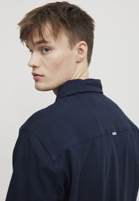 Chemise bleu marine avec un col classique et des coutures visibles. Présente une petite étiquette à l'arrière et un tissu lisse et texturé.