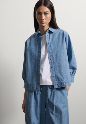 Frau mit langen dunklen Haaren trägt ein lockeres blaues Hemd über einem weißen T-Shirt und passenden blauen Hosen, steht vor einem einfarbigen Hintergrund.