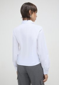 Camicia bianca a maniche lunghe con colletto, caratterizzata da una texture liscia. Polsini con bottoni e un design aderente. Indossata con pantaloni grigi a righe.